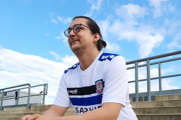 Junger Mann in einem Sporttrikot sitzt auf einer Tribüne unter blauem Himmel.