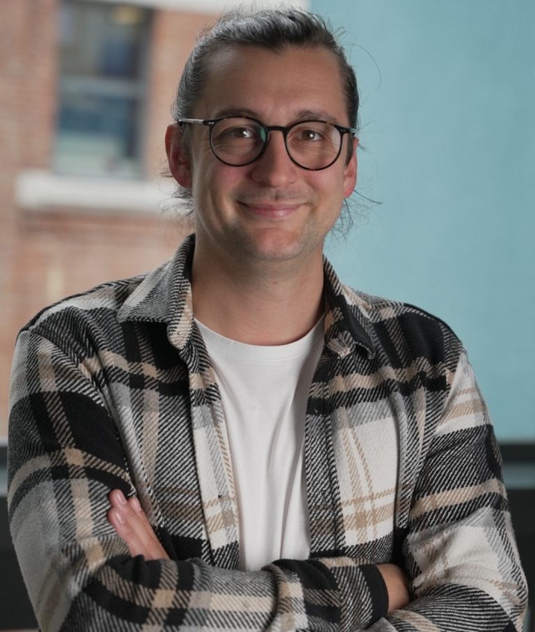 Junger Mann mit Brille und lässiger Kleidung, lächelt und steht mit verschränkten Armen.