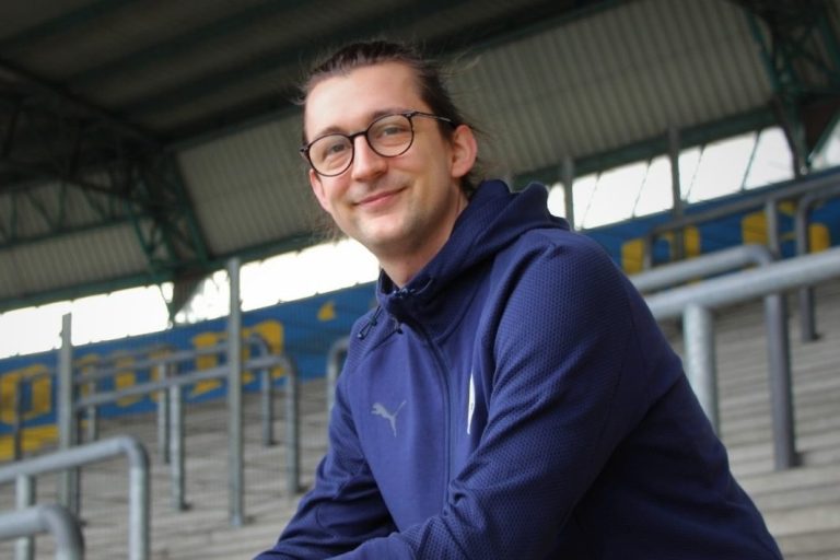 Lächelnder Mann in blauer Jacke sitzt auf einer Tribüne im Stadion.