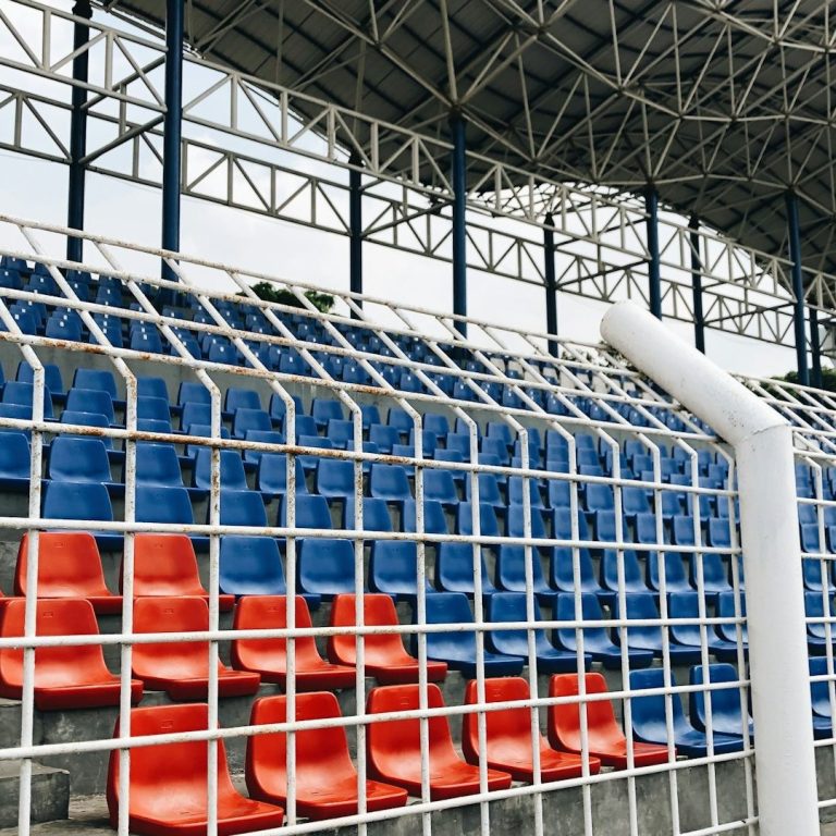 Leere Stadiontribüne mit blauen und roten Sitzen, umgeben von einem Gitter.